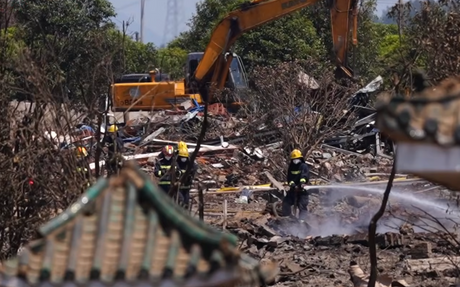 Aftermath of the Liuyang fireworks plant explosion: firefighters working in debris, with a yellow excavator behind and a Chinese tile roof in the foreground. 5 May 2026.