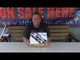 Presenter holding Tropical Storm 40-shot firework cake by Evolution Fireworks in front of a promotional banner at a fireworks retail store.